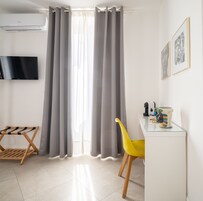 Una habitación moderna con televisión de pantalla plana, cortinas grises, una silla de madera, una silla amarilla y una mesita con objetos decorativos.
