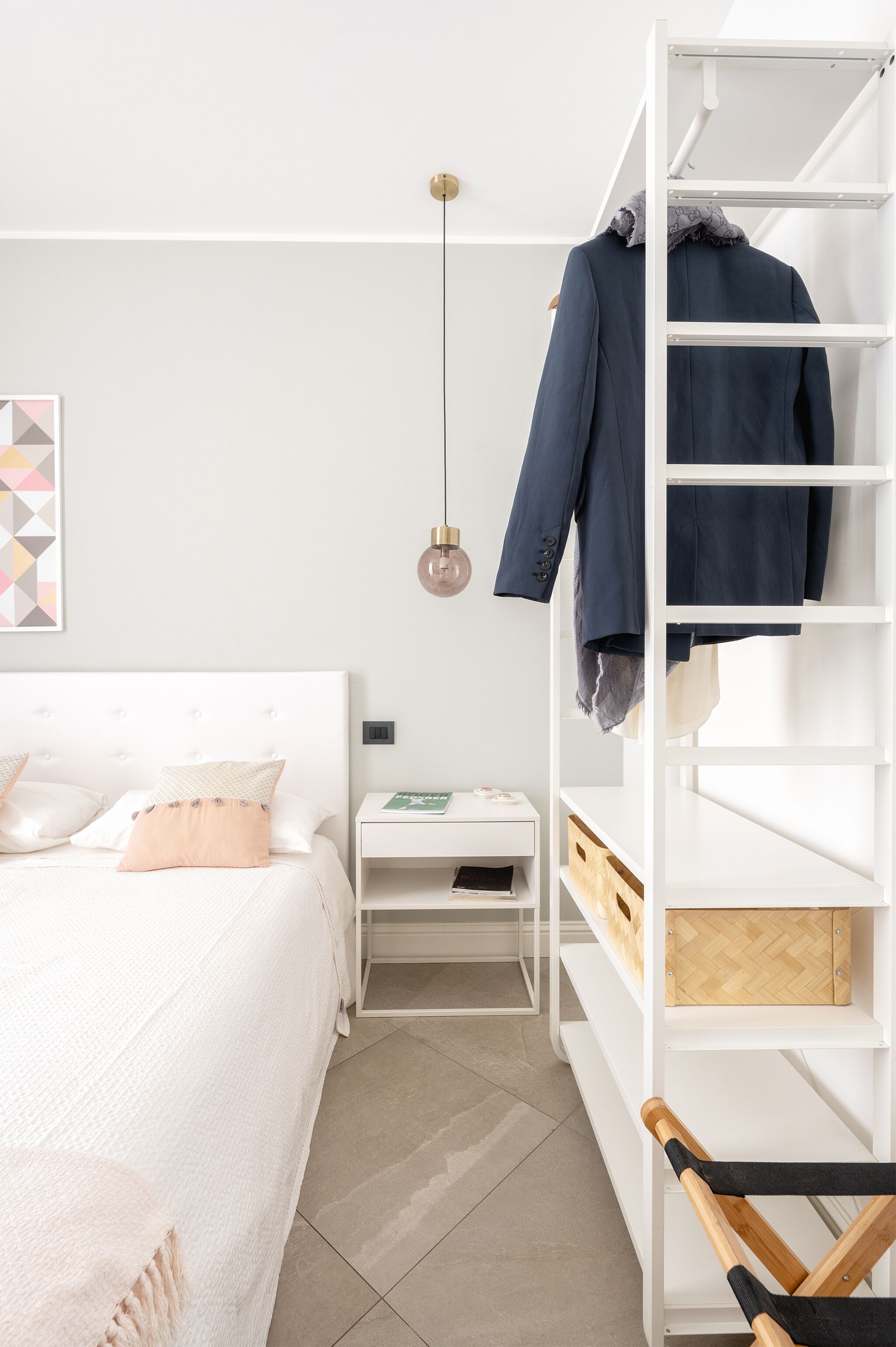 Una camera da letto ordinata e accogliente, con un letto, un comodino bianco, una scaffalatura bianca e una lampada a sospensione.