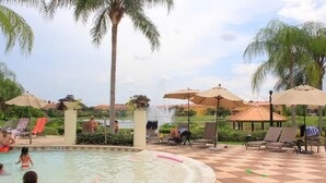 A pool area with lounge chairs and umbrellas, surrounded by palm trees and a view of houses in the background. - Ly86465 - Encantada Resort - 2 Bed 2 Baths Townhome (Kissimmee)