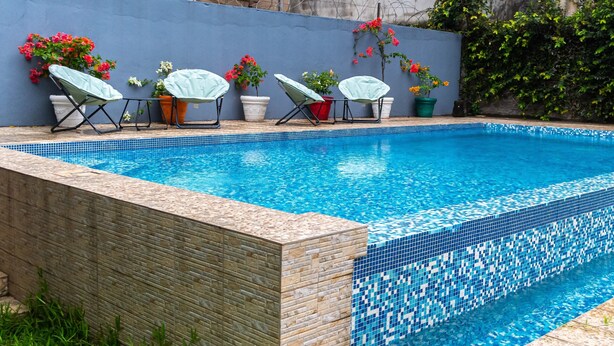 A rectangular swimming pool with blue mosaic tiles, surrounded by a stone wall and potted plants. - VMD Aparthôtel (Abidjan)
