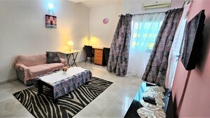 A living room with a sofa, a coffee table, a lamp, a TV, and a window with curtains. - VMD Aparthôtel (Abidjan)