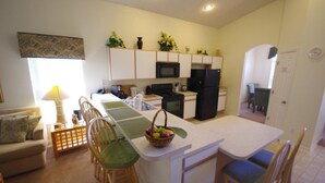 Una cocina funcional con gabinetes blancos, microondas, estufa y refrigerador. Hay un comedor con mesa y sillas.