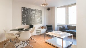 A modern living room with a dining area, a large black and white cityscape artwork on the wall, a black leather chair, a grey sofa, and a glass coffee table. - App De Panne (De Panne)