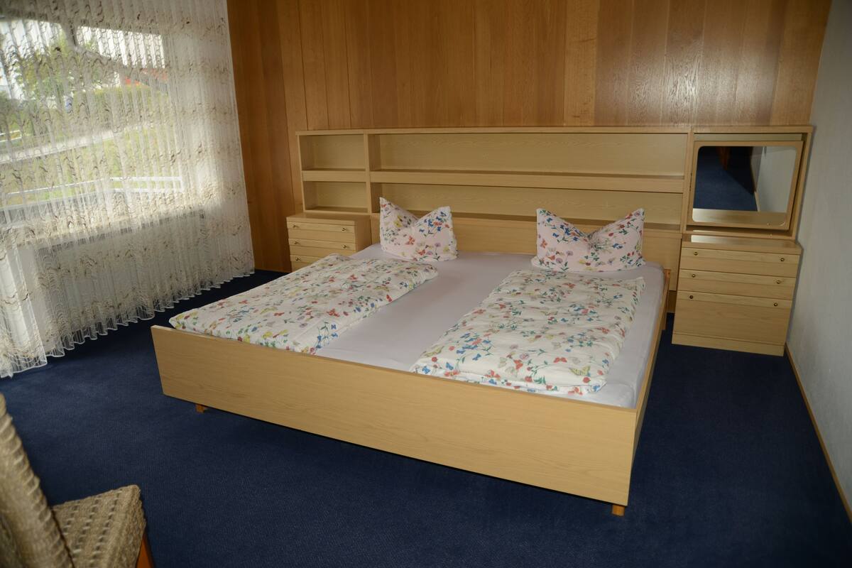 Une chambre à coucher avec un lit en bois, une literie à motifs floraux, un miroir et une fenêtre ornée de rideaux en dentelle.