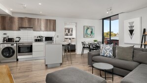 A modern kitchen with white cabinets, a built-in oven, and a washing machine. There is a dining area with a table and chairs, and a gray sofa in the living room. - Greenwich 602 (Cape Town)