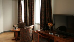 A hotel room with a wooden desk, two patterned armchairs, a television, and a window with curtains.