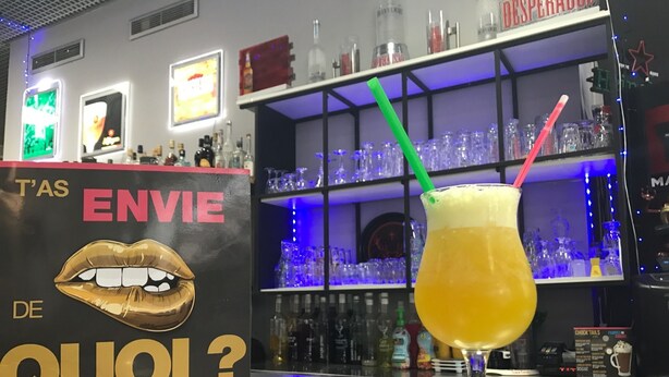 A bar area with a drink in a glass, a sign with lips and text, and shelves with bottles and glasses.