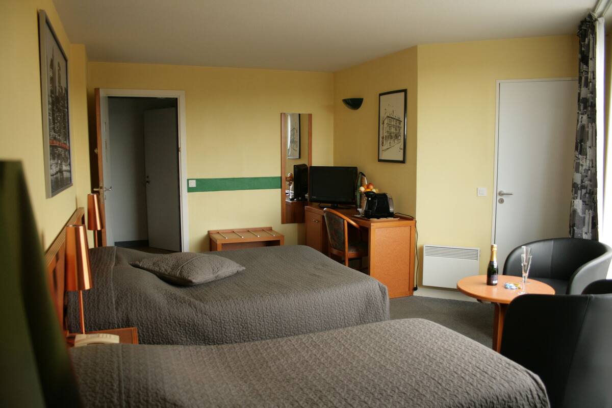 Une chambre d’hôtel avec deux lits, un bureau avec un ordinateur, une chaise, une petite table avec une bouteille et un verre, et une porte.