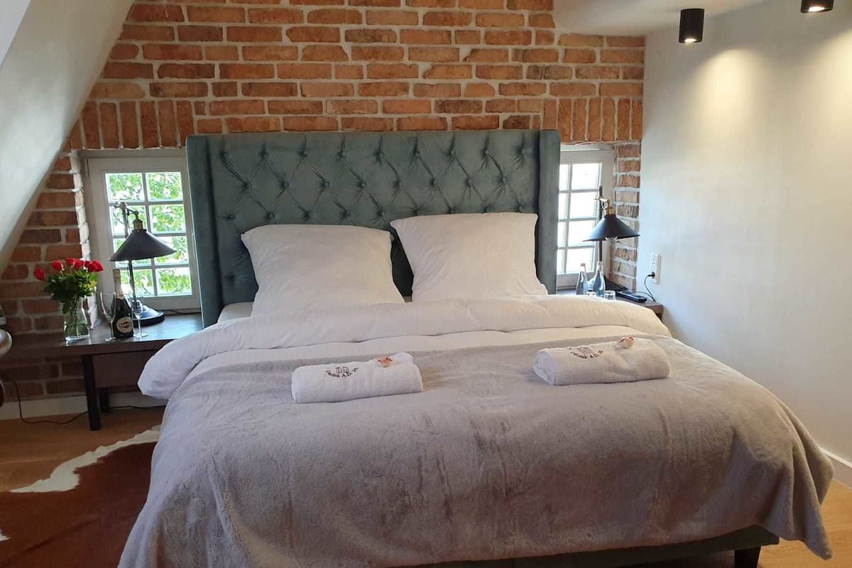 A bedroom with a brick wall, a tufted headboard, a large bed with white linens, and a cowhide rug.