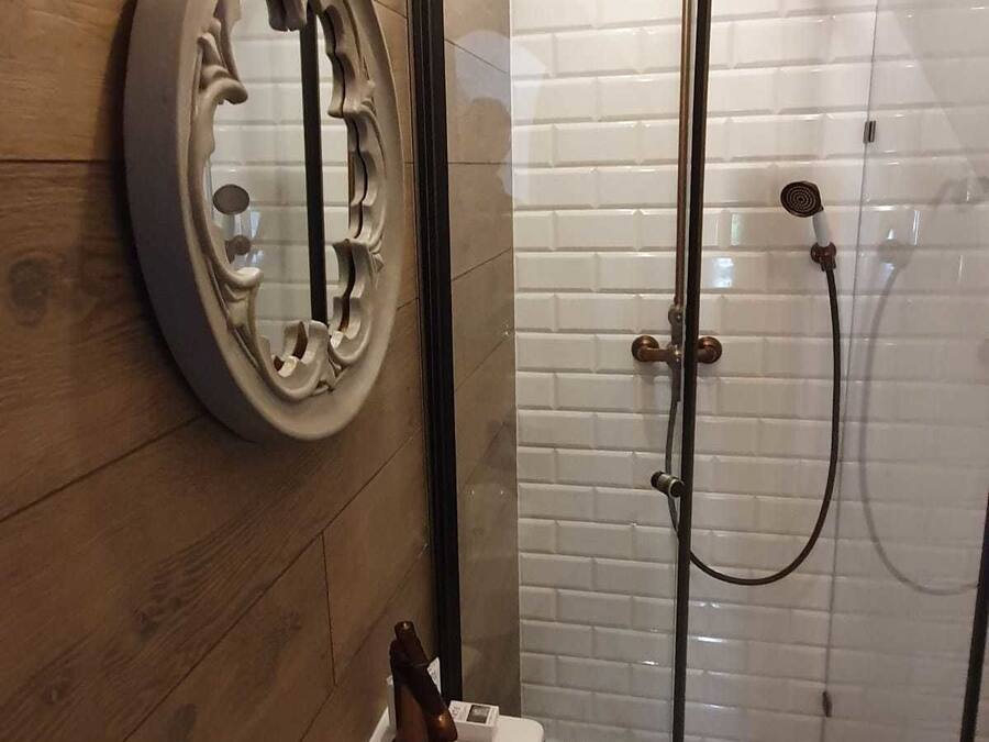Un baño con piso de madera, inodoro blanco, ducha con puerta de vidrio y espejo con marco decorativo.