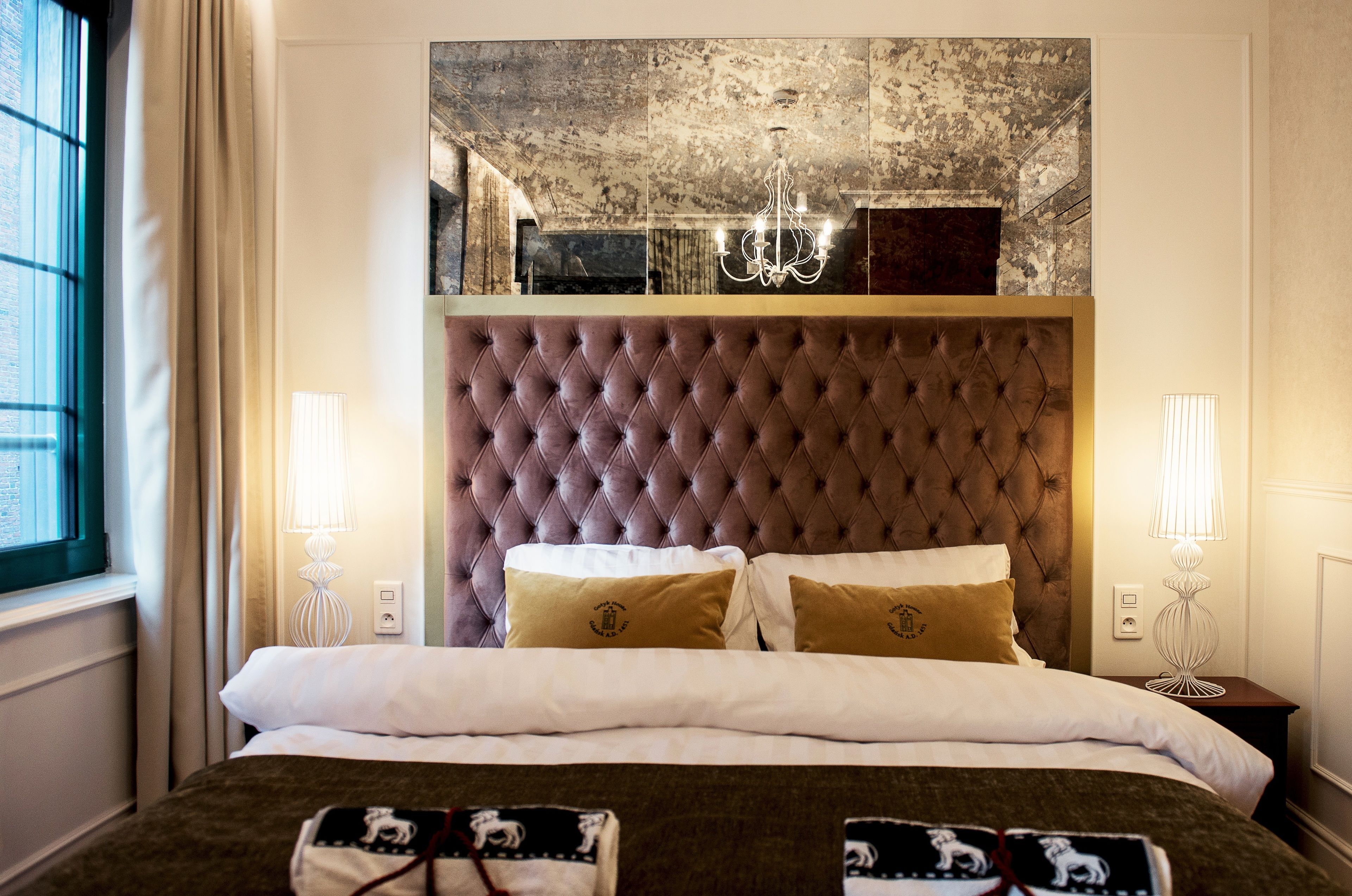 A neatly made bed with a tufted headboard, two bedside lamps, and a large window with curtains.