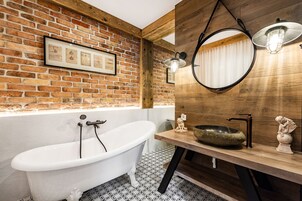 A bathroom with a white clawfoot bathtub, a wooden vanity with a round mirror, and a brick wall with framed pictures.