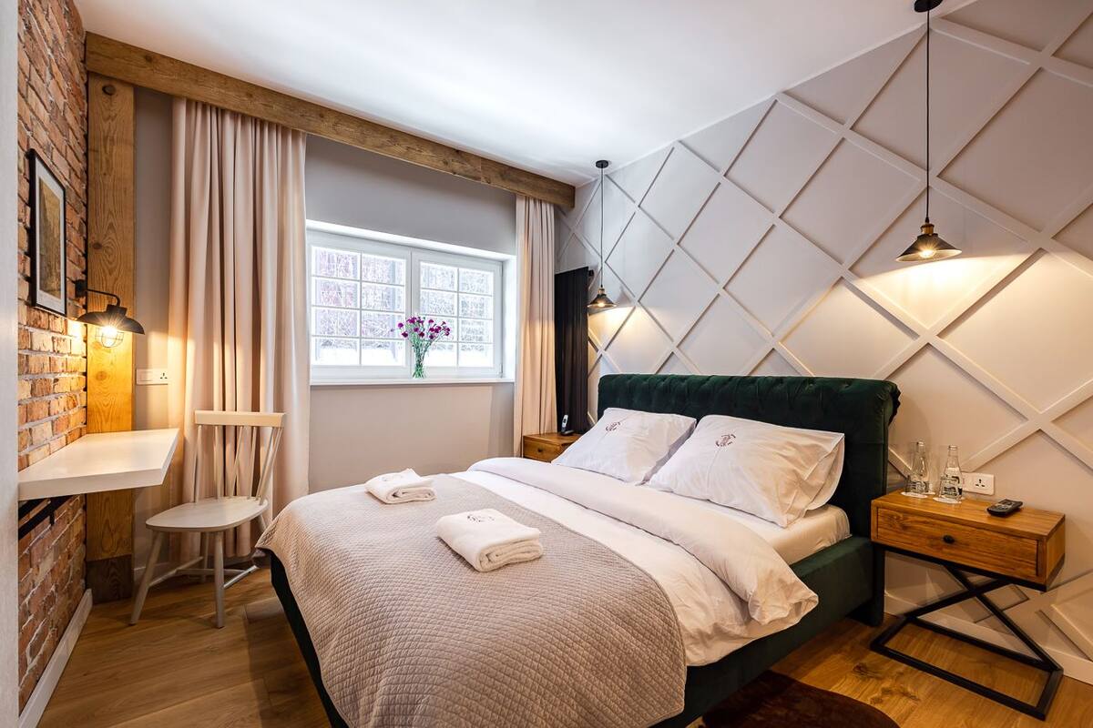 A bedroom with a bed, a desk, a chair, a window with curtains, and a wall with a geometric pattern.