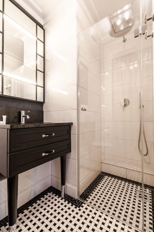 Un baño moderno con piso de baldosas blanco y negro, cabina de ducha de vidrio y tocador con espejo y lavamanos.