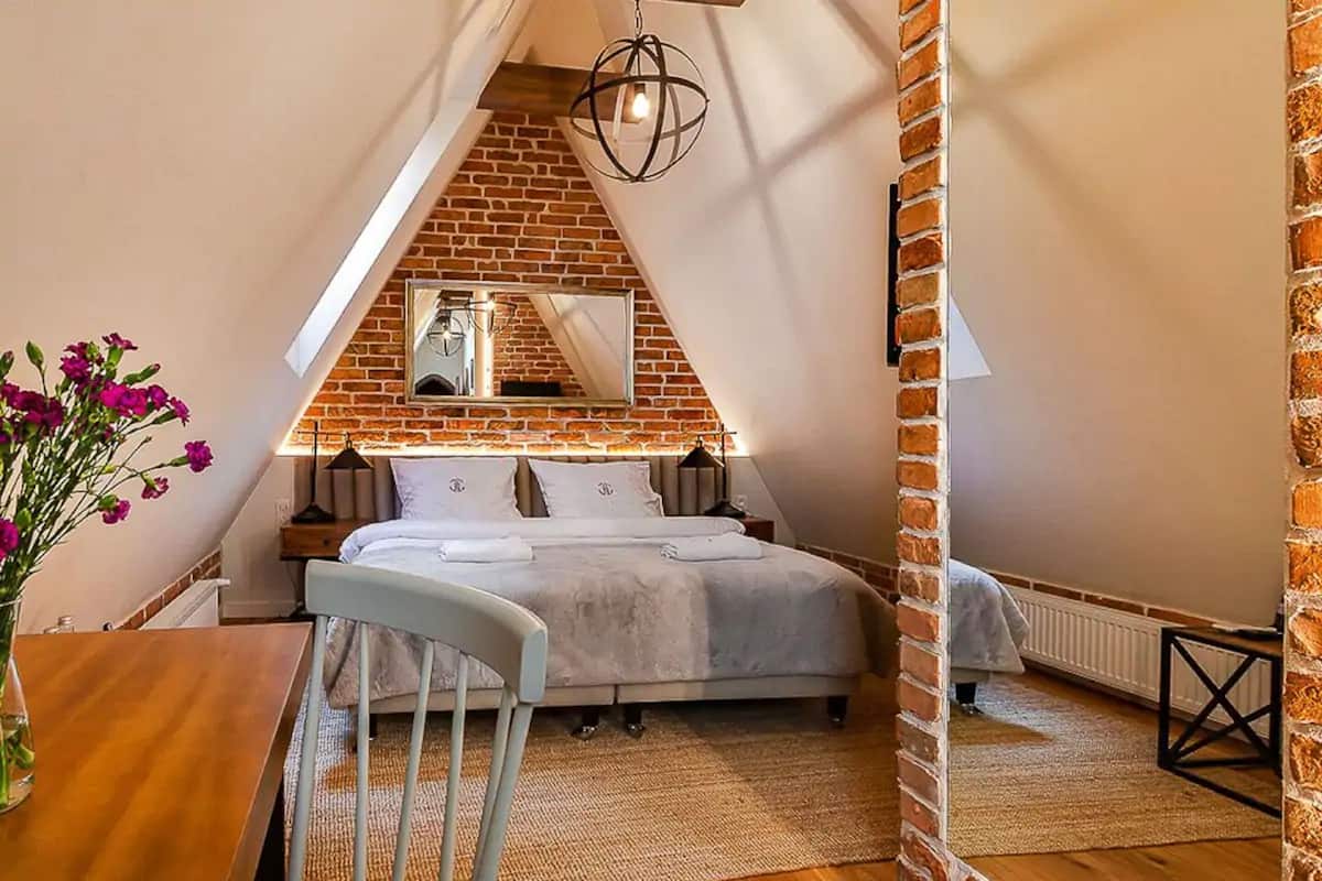 A bedroom with a sloped ceiling, a brick wall, a bed, a chair, a table, and a vase of flowers.