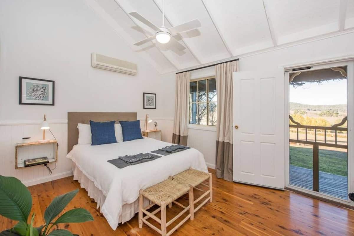 A bedroom with a bed, bedside tables, a ceiling fan, a window with curtains, and a view of the outdoors.