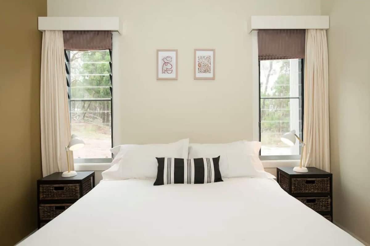 A neatly made bed in a room with two windows, each with a curtain and a framed picture on the wall.