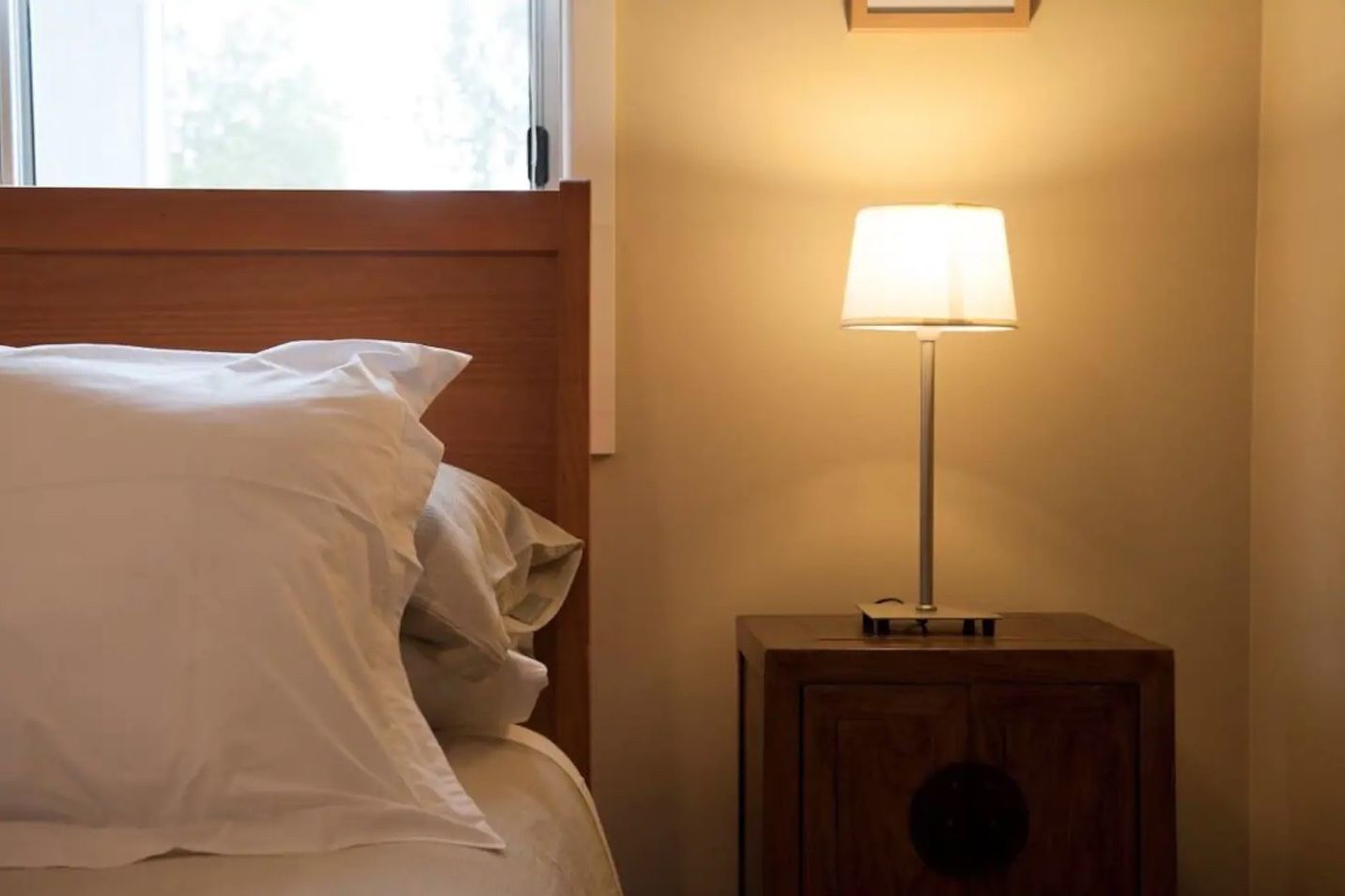 A neatly made bed with white linens, a bedside table with a lamp, and a window with blinds.