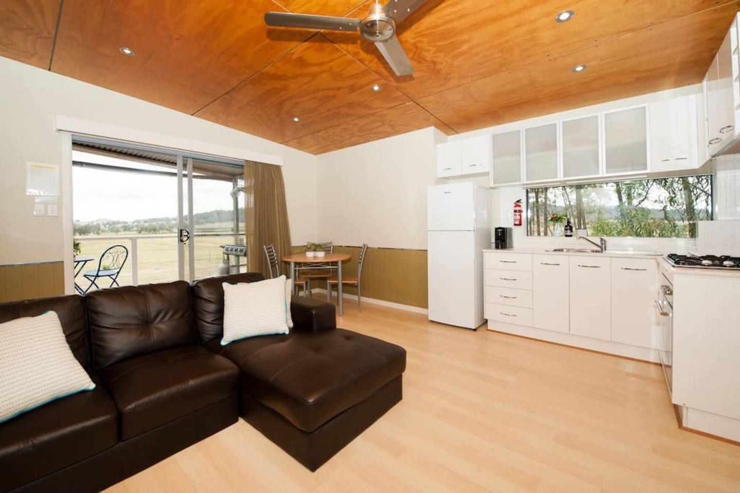 Una sala de estar moderna con un sofá de cuero marrón, un comedor con mesa y sillas, y una cocina con armarios blancos y estufa.