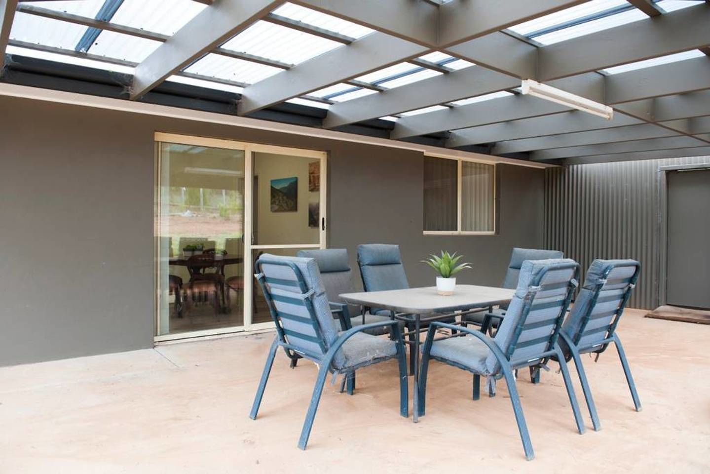 Un patio cubierto con una mesa redonda y seis sillas, una planta en maceta y un cerramiento de vidrio corredizo que da a un espacio interior.