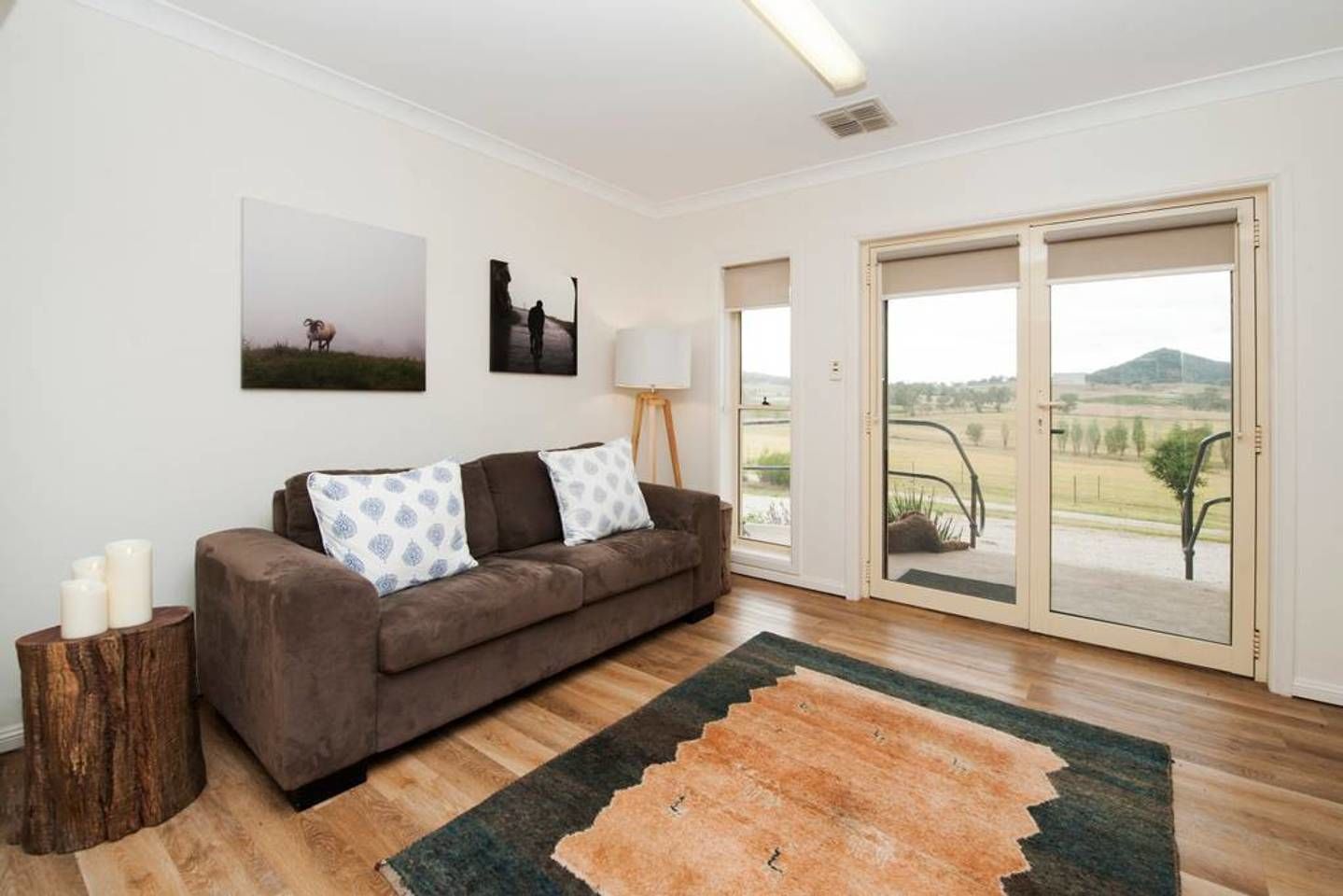 A modern living room with a brown sofa, two patterned cushions, a wooden side table with candles, a large window with a view of the countryside, and a floor-to-ceiling sliding door.