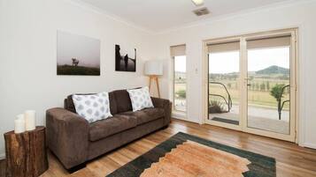 A modern living room with a brown sofa, two patterned cushions, a wooden side table with candles, a large window with a view of the countryside, and a floor-to-ceiling sliding door.