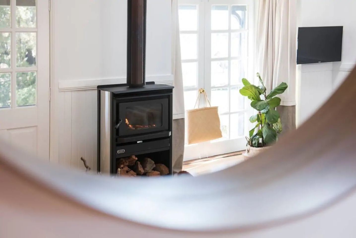 Una habitación con estufa de leña, ventana con cortinas y una planta en maceta.