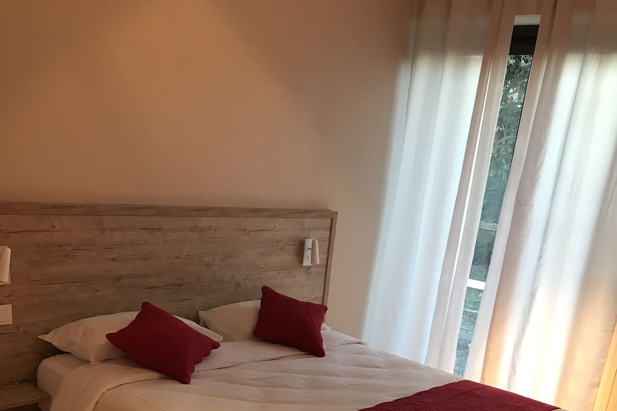 Un lit bien fait, avec du linge de lit blanc et des touches de rouge, une tête de lit en bois et une vue sur la verdure à travers des rideaux transparents.