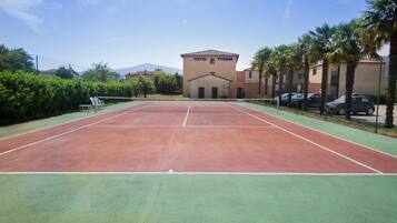 Una cancha de tenis con una red, rodeada por una cerca y palmeras. Al fondo, se observa un edificio con la leyenda 'HOTEL PYRENE'.