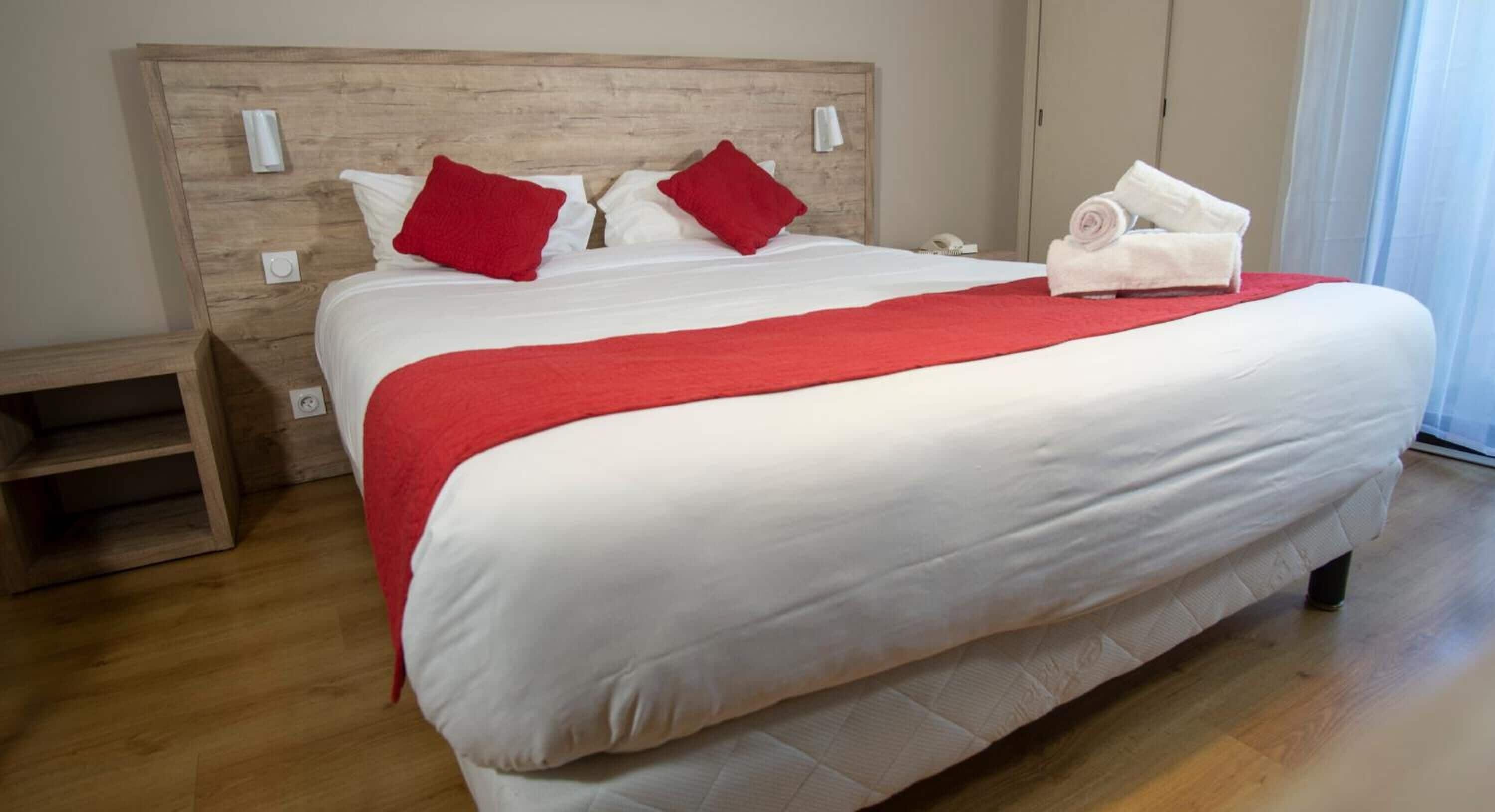 Un lit bien fait, avec une literie blanche et rouge, une tête de lit en bois et un plaid rouge.