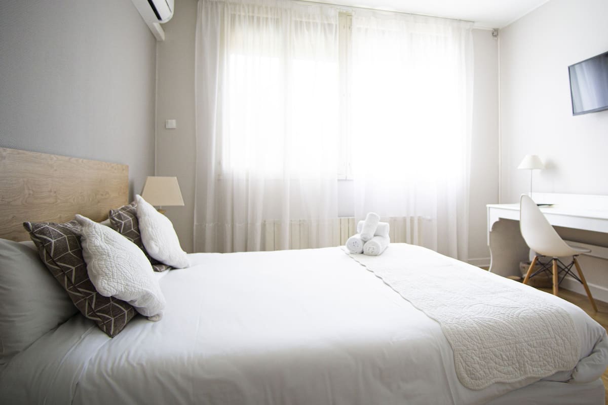 Une chambre d’hôtel équipée d’un lit, d’un bureau, d’une chaise, d’une télévision et d’une fenêtre avec des rideaux légers.