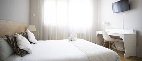 A hotel room with a bed, a desk, a chair, a television, and a window with sheer curtains.