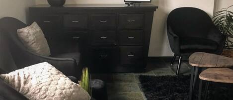 Una sala de estar con un sofá negro, un sillón negro, una mesita redonda, un tapete negro, un aparador con cajones, un cuadro enmarcado y un jarrón con flores.