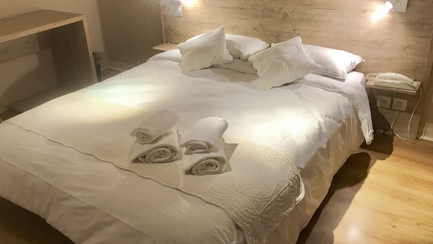 Un lit bien fait, avec du linge de lit blanc et des serviettes pliées, une tête de lit en bois et des lampes fixées au mur, ainsi qu’un téléphone sur la table de chevet.