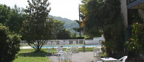 Un patio al aire libre con sillas y mesas de plástico blanco, rodeado de vegetación y vistas a una piscina.