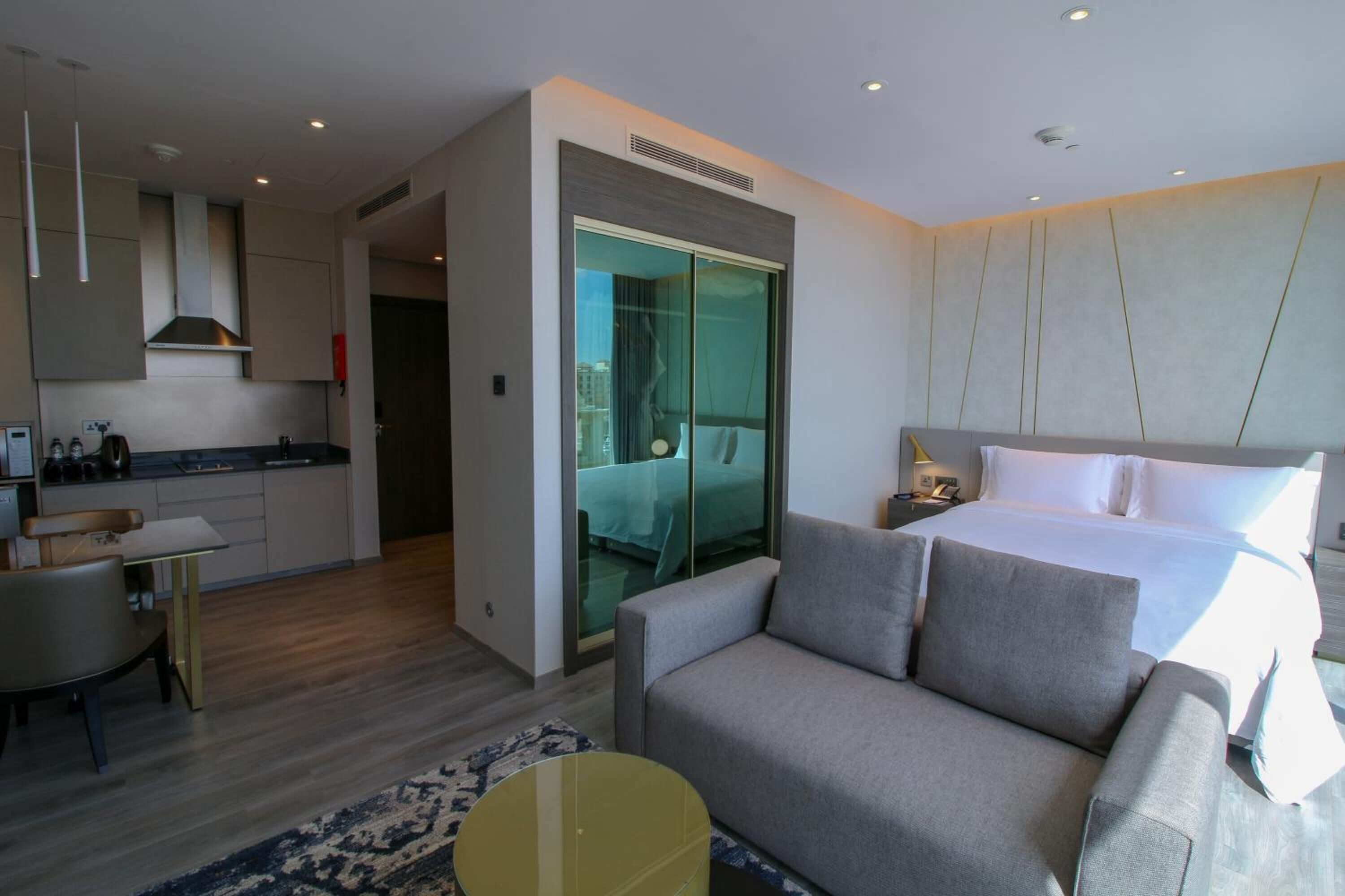 A modern hotel room with a sofa, a bed, a glass door leading to a balcony, and a kitchenette.