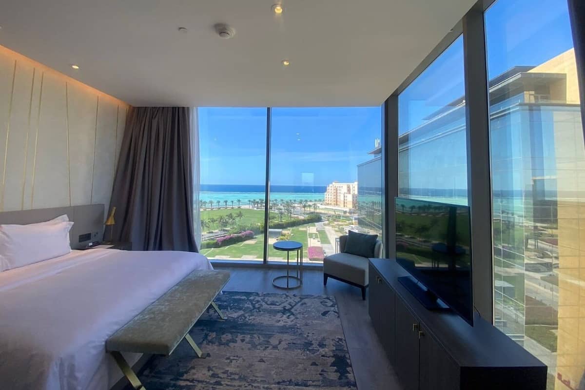 A hotel room with a large bed, a small seating area, a TV, and a balcony with a view of the ocean and buildings.
