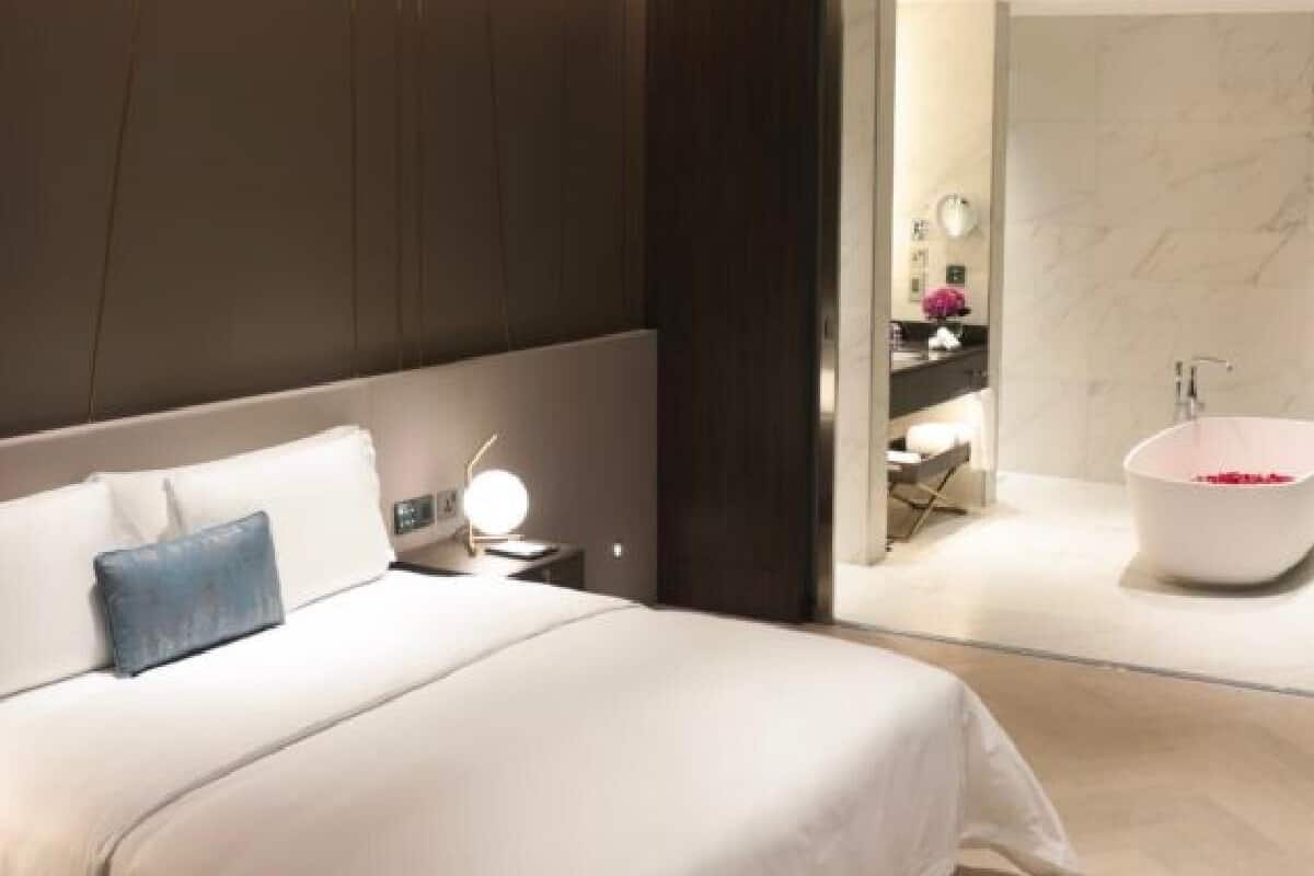 A modern bedroom with a large bed, a bedside table with a lamp, and a view into a bathroom with a bathtub.