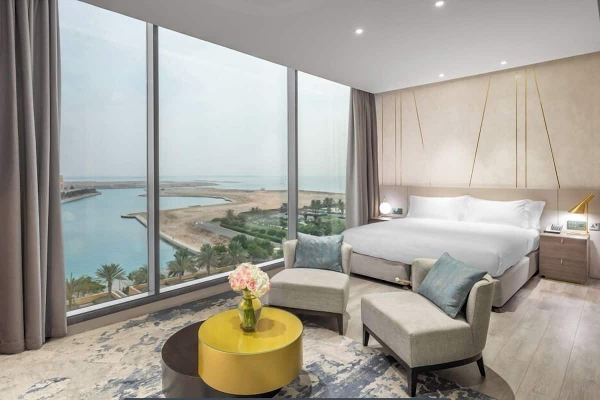 A modern hotel room with a large bed, a sofa, a small table, and a view of the coast.