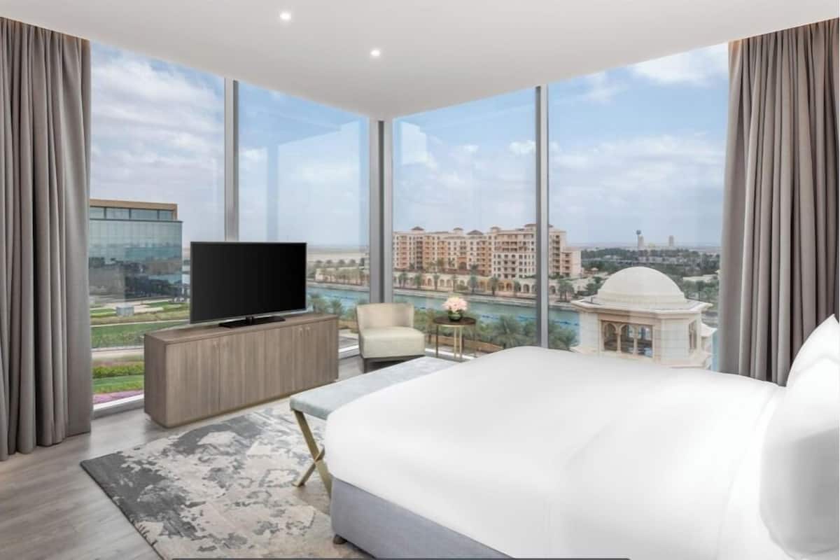 A modern hotel room with a large bed, a flat-screen TV, a small table with a lamp, and a view of a cityscape through large windows.