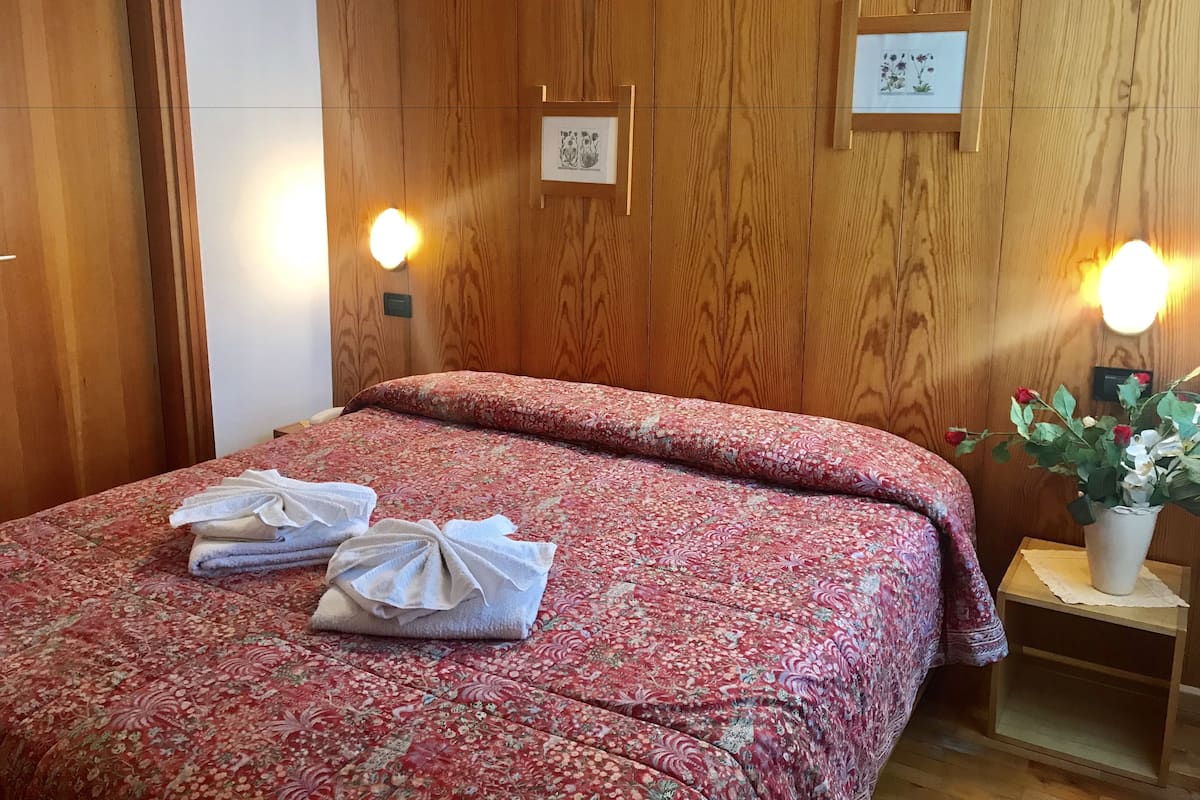 A neatly made bed with a floral bedspread, towels, and a small bedside table with a vase of flowers.