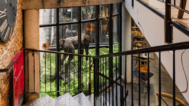 Un interior de varios niveles con una escalera, un balcón con vistas a un área verde exterior y una rústica pared de piedra con relojes.