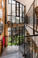 Un interior de varios niveles con una escalera, un balcón con vistas a un área verde exterior y una rústica pared de piedra con relojes.