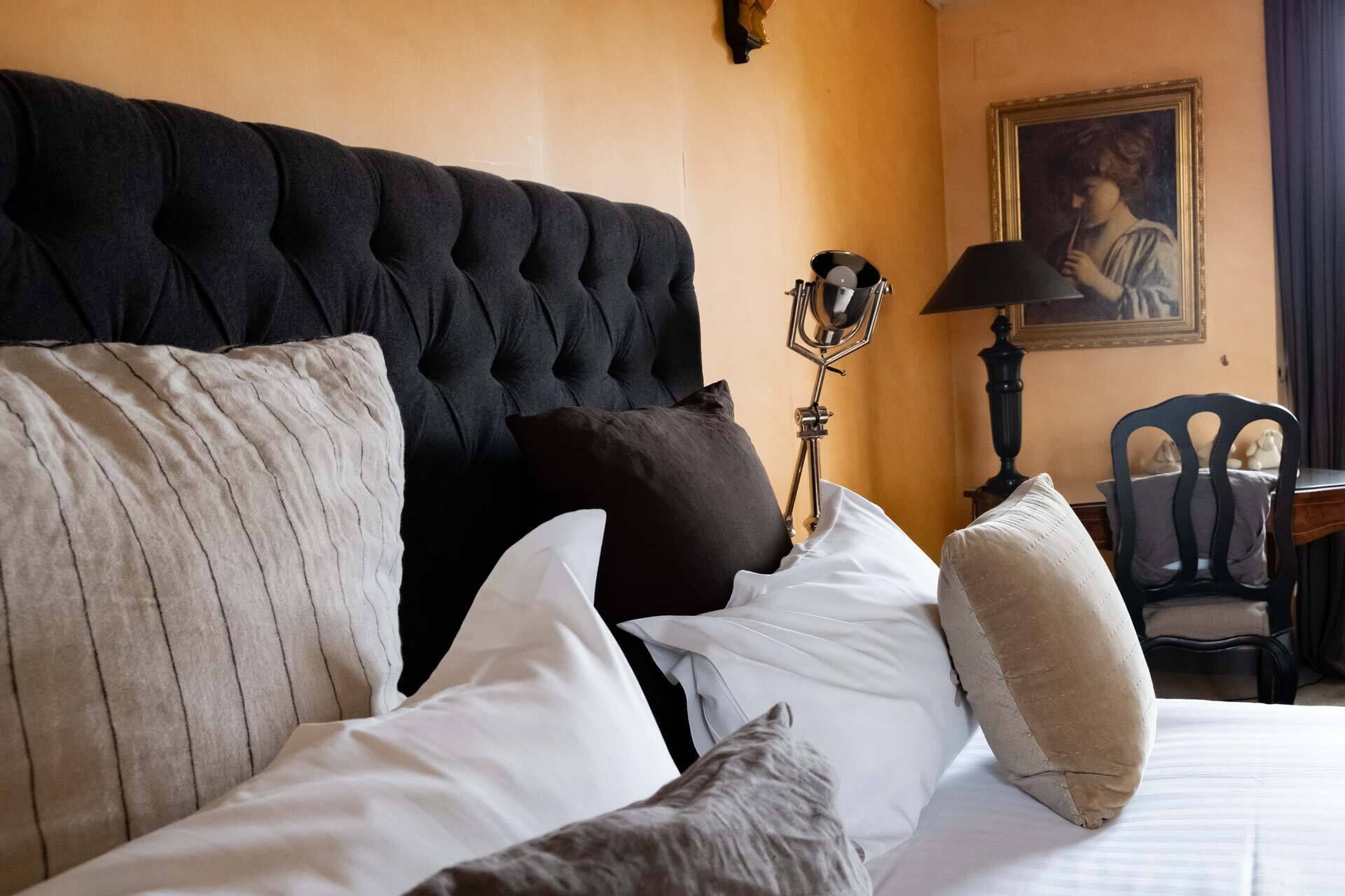 A neatly made bed with multiple pillows and a tufted headboard. A lamp and a chair are visible in the background.