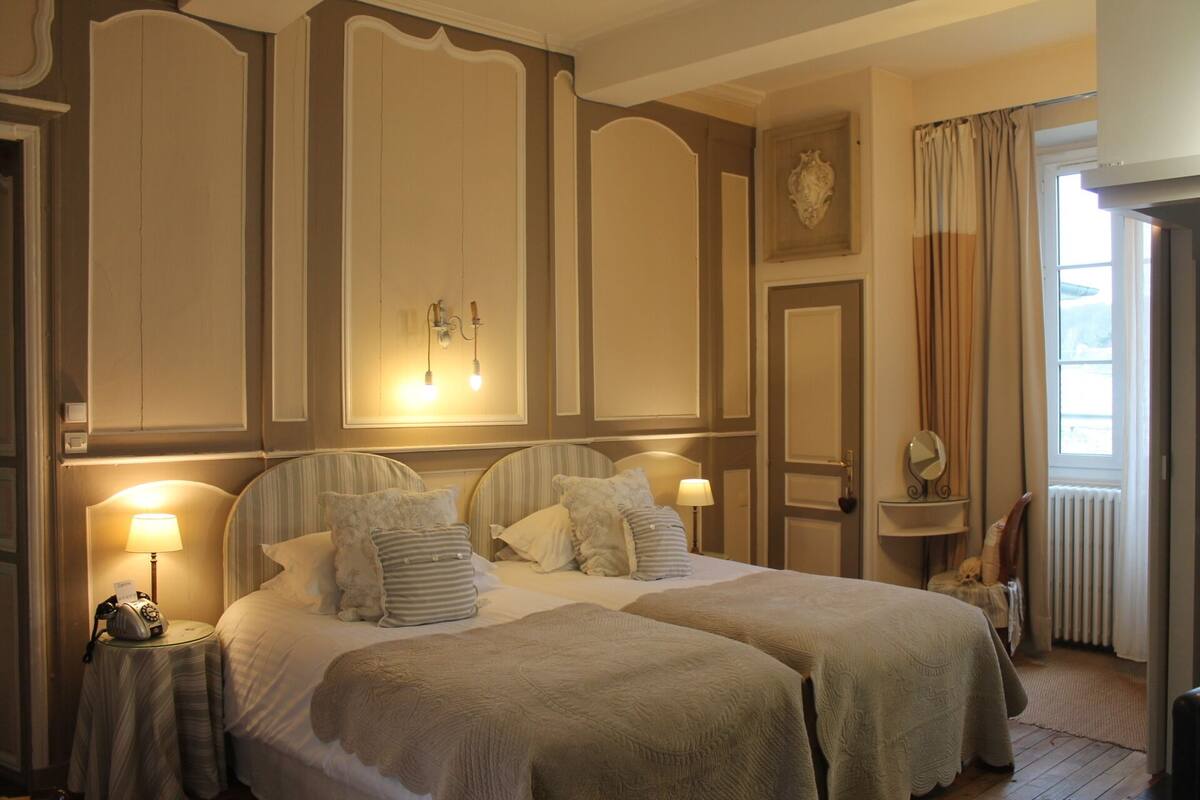 A hotel room with two beds, a small table with a lamp, a clock, and a mirror on the wall.