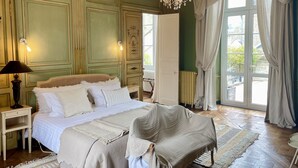 Un dormitorio con una cama grande, una mesita de noche, una silla, una araña y vistas a través de una ventana con cortinas.
