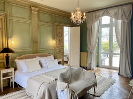 Un dormitorio con una cama grande, una mesita de noche, una silla, una araña y vistas a través de una ventana con cortinas.