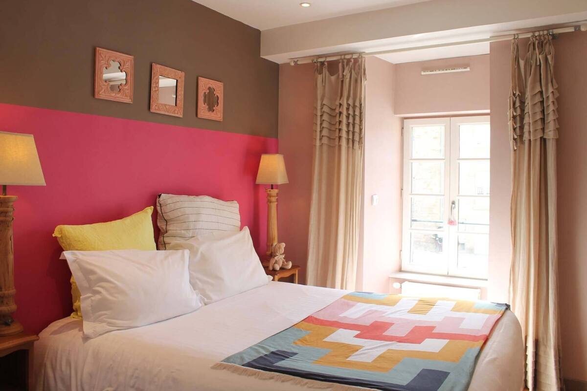 A bed with a colorful quilt, two bedside lamps, and a window with curtains.