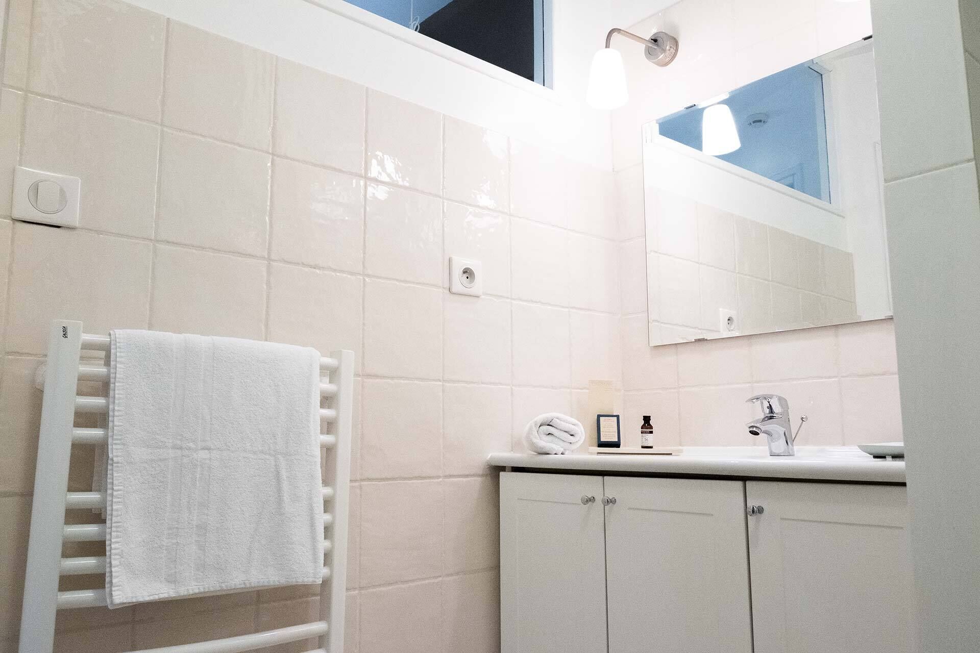 Un baño con paredes de azulejos blancos, una toalla blanca en un perchero, un espejo, un lavabo con grifo y un armario.