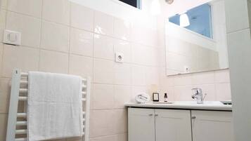 Un baño con paredes de azulejos blancos, una toalla blanca en un perchero, un espejo, un lavabo con grifo y un armario.